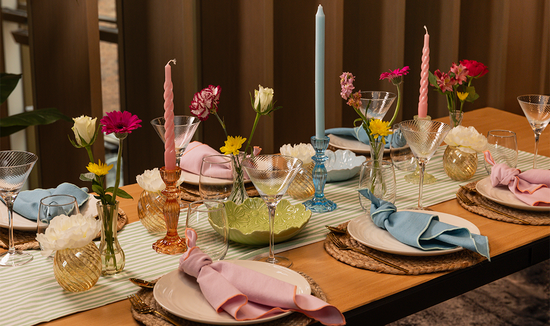 table setting with pink and blue napkins knotted on plates which are on rattan placemats. Green stripe table runner lines the centre of the table.