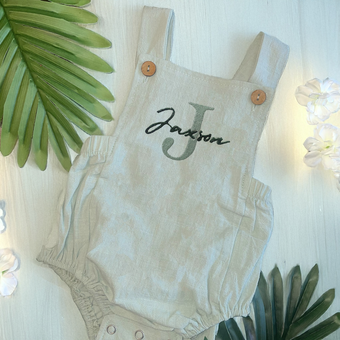 A beige personalised romper on a pink hanger. A pair of baby shoes with a cream ribbon. 